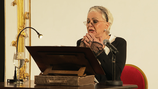 Eine ältere Dame steht vor einem Lesepult und spricht bedächtig in ein Mikrofon.