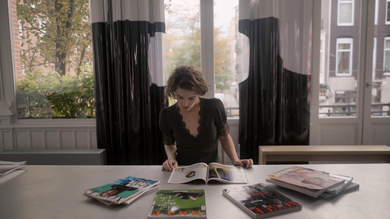 Die Soziologin Sylvia Holla sitzt am Tisch vor einer Fensterfront und blättert Modemagazine durch. 