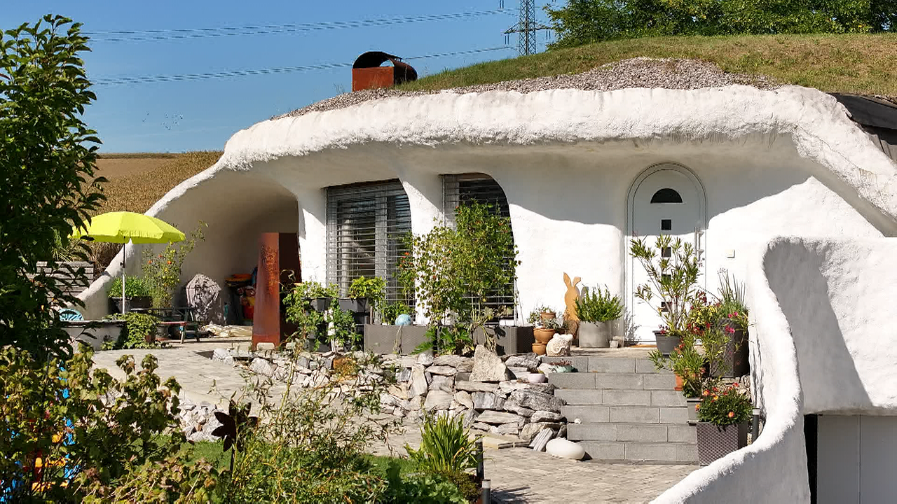 Kalkweißes, rundes Haus mit bewachsenem Dach.