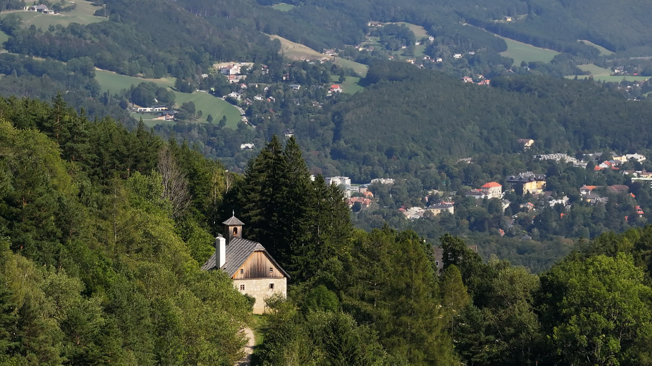 Ein kleines Haus mit Glockenturm, umgeben von Wald in einer bergigen Gegend, aus der Vogelperspektive.