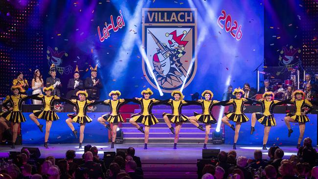 © ORF/Peter Krivograd Die "Villacher Fasching" in gelb‑schwarzen Kostümen tanzt in einer Reihe synchron vor dem Villach‑Wappen und dem Schriftzug „Lei‑Lei 2026“.