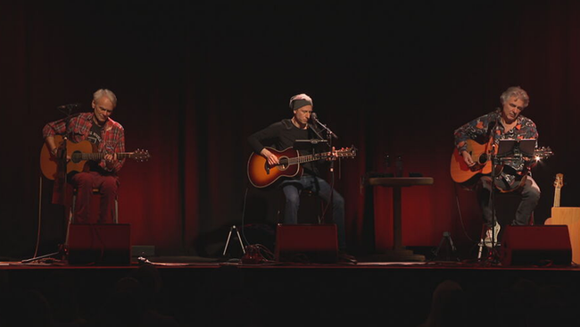 Die drei Musiker sitzen mit Gitarren in der Hand auf der Bühne, roter Vorhang im Hintergrund, intime Lichtstimmung
