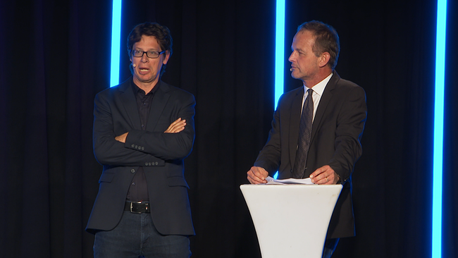 Zwei Männer mit Mikrofonen stehen auf einer Bühne.