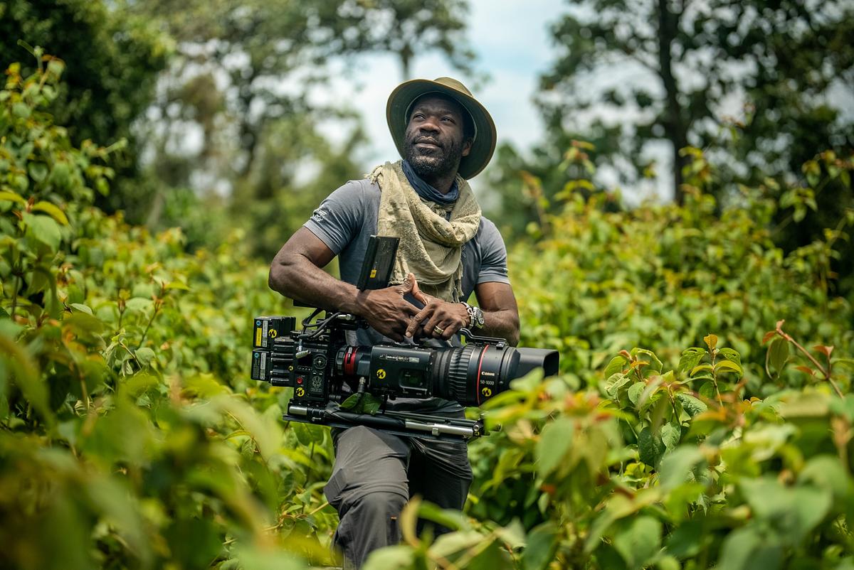Der Kameramann hat seine Kamera auf seinem rechten Oberschenkel abgestützt. Er ist von grünen Büschen und Bäumen umgeben.