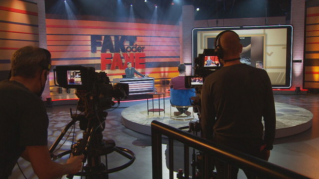 Hinter den Kulissen der ORF Sendung Fakt oder Fake. Zwei Kameramänner stehen während der Aufzeichung im Studio. Der Moderator und seine Gäste sind zu sehen. 