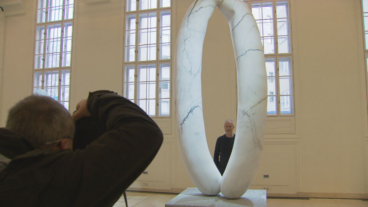 Erwin Wurm steht hinter einer Würstel Skulptur aus Marmor und wird dabei fotografiert 