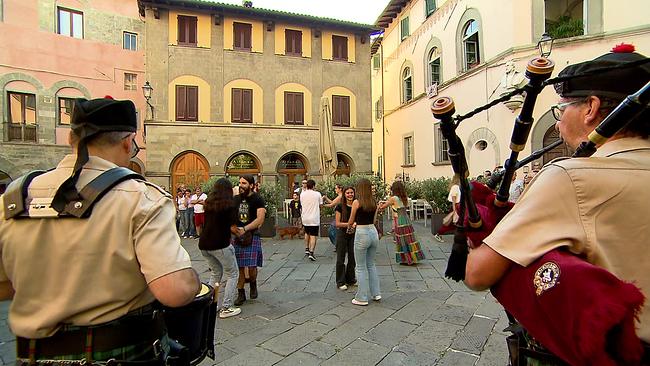 Platz in Barga umgeben von alten Häusern, junge Menschen tanzen paarweise, im Vordergrund sieht man 2 Dudelsackspieler von hinten
