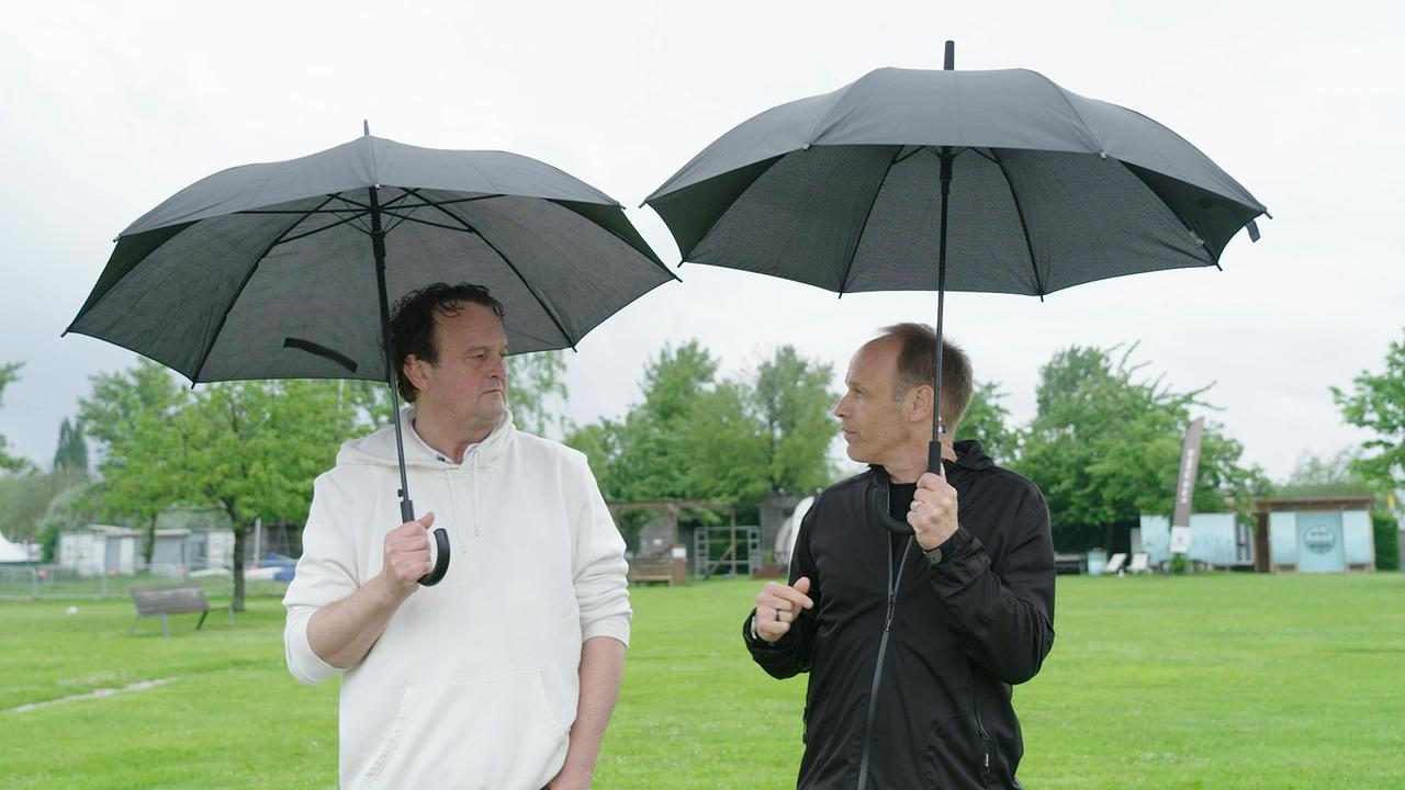 Hanno Settele steht neben Markus Wadsak. Beide halten einen Regenschirm in der Hand und warten auf den erlösenden Regen.