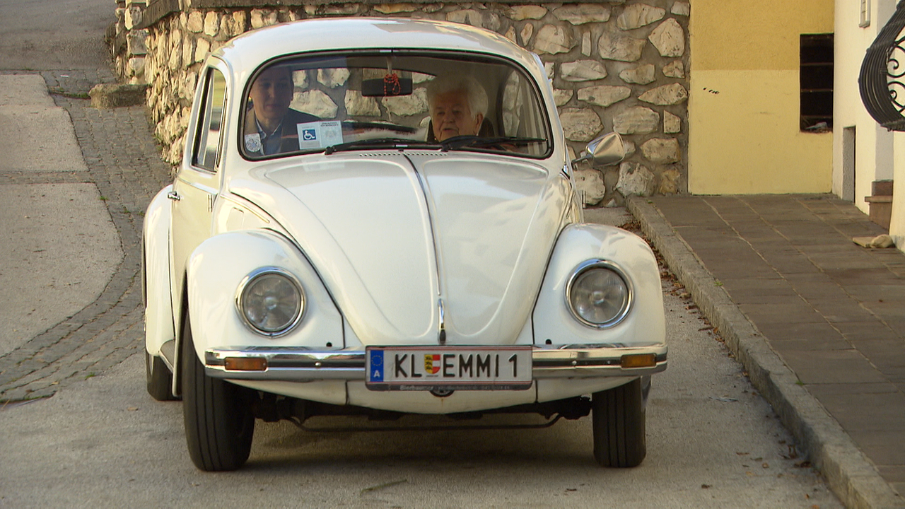 Frau M. in einem weißen alten VW-Käfer auf der Straße. 