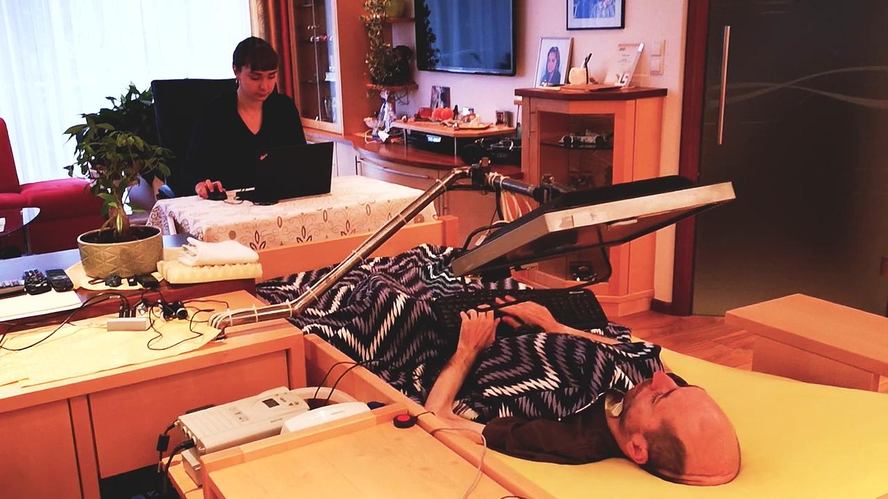 Herr W. ist zu sehen, wie er im Bett liegend mit seinem speziellen Equipment im Homeoffice arbeitet,  trotz seiner Erkrankung
