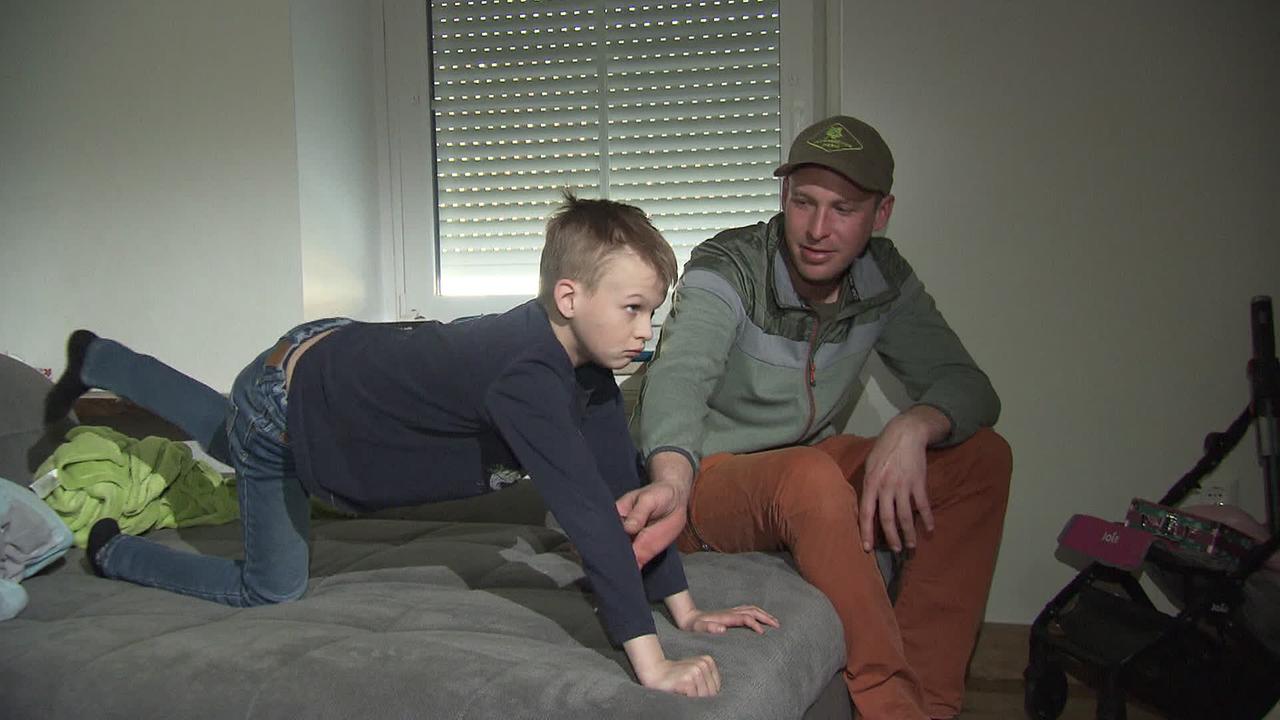 Ein Bub versucht Bewegungsübungen am Bett zu machen. Sein Vater gibt ihm Hilfestellung.