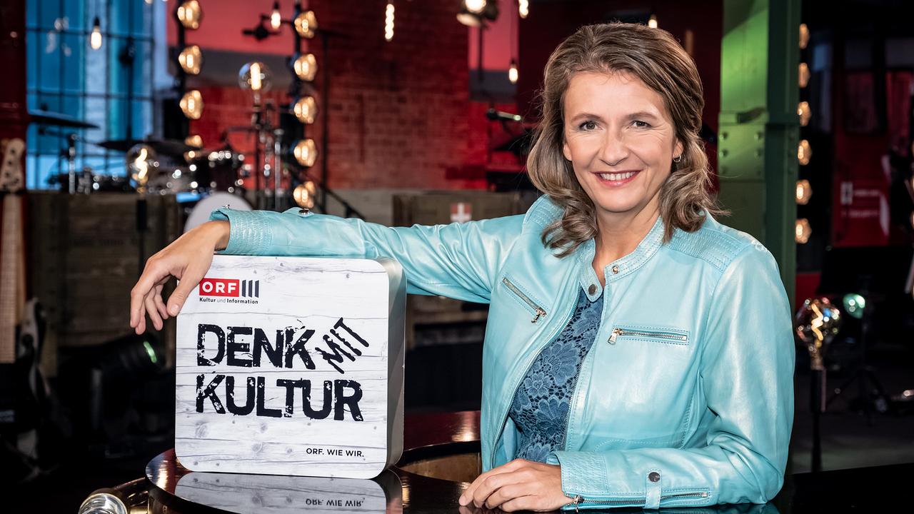 Birgit Denk befindet sich im Verkehrsmuseum Remise - sie trägt eine türkise Lederjacke und stützt sich auf einen Leuchtkasten auf dem der Sendungstitel DENK mit KULTUR steht