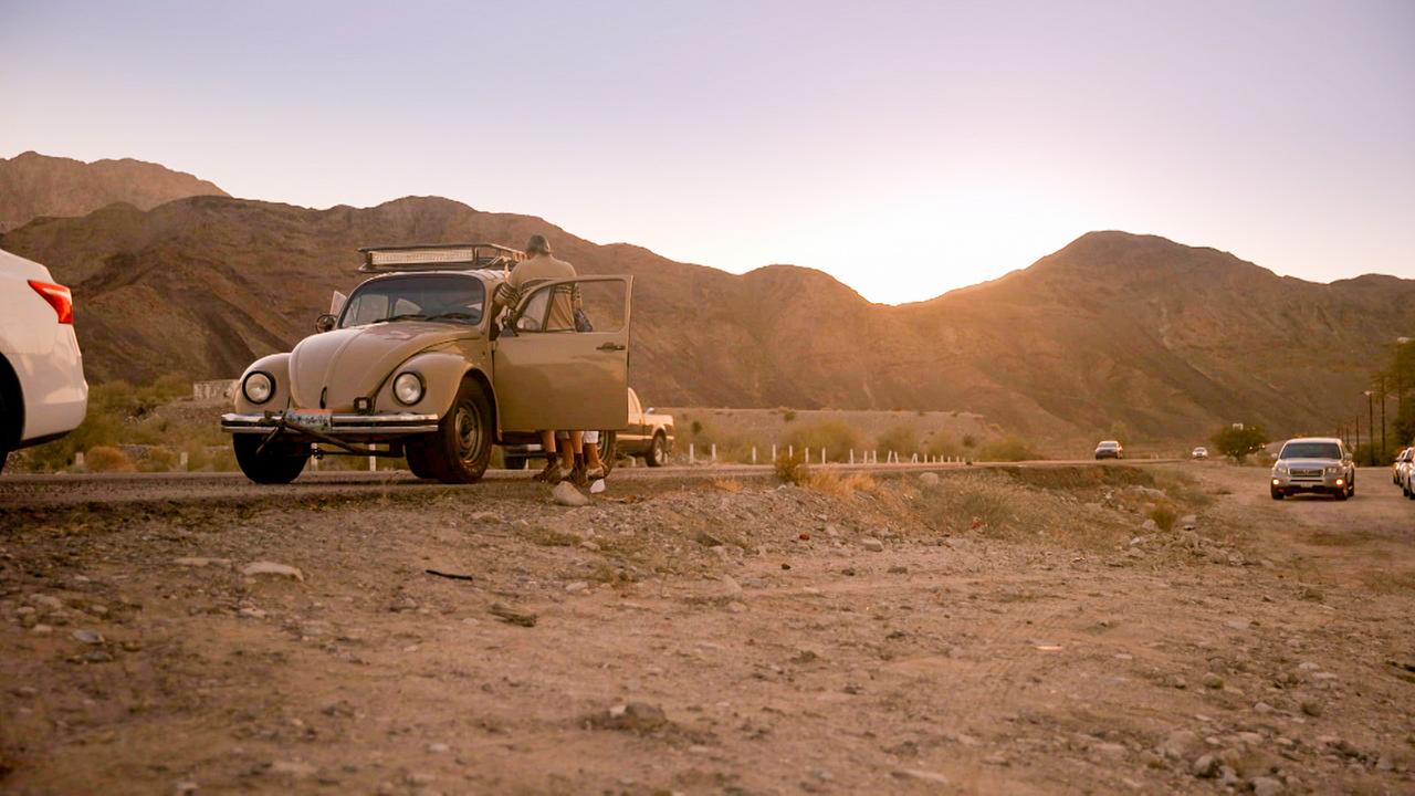 Am Rande einer Straße im trockenen Baja California steht ein alter Käfer, weitere Autos im Hintergrund fahren auf der Straße.