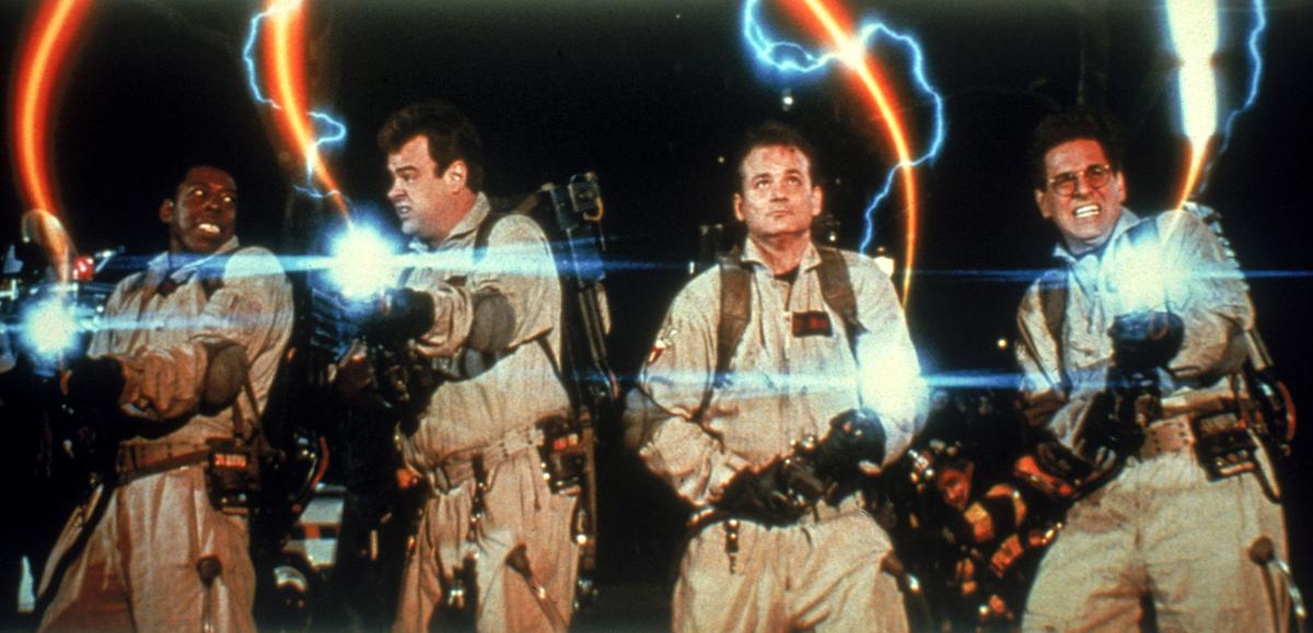 Vier Ghostbusters mit ihrer Ausrüstung in beigefarbenen Overalls mit, um sie herum blaue Blitze und orangefarbe Strahlen