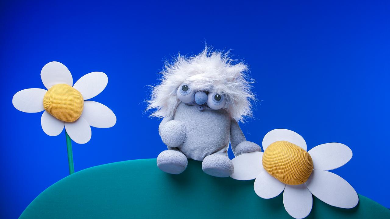 die graue Puppe von Lollobien sitzt auf einem Hügel neben zwei weißen Blümchen.