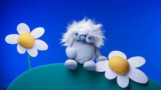 die graue Puppe von Lollobien sitzt auf einem Hügel neben zwei weißen Blümchen.
