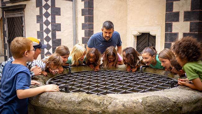 © Kultur-Betriebe Burgenland Eine Kindergruppe und ihr Guide haben sich um einen alten Burgbrunnen versammelt und schauen gespannt durch das Gitter nach unten.