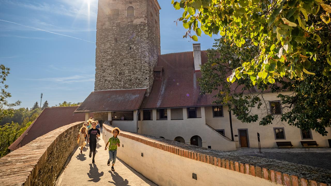 Ein paar Kinder laufen aus der Burg Schlaining heraus. Hinter dem Burgturm blinzelt die Sonne hervor. 