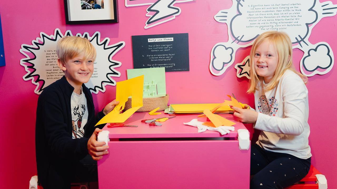 Im Bild: Ein Bub und ein Mädchen sitzen vor einer rosa Wand an einem rosa Tisch und schneiden gelbes Papier aus. 