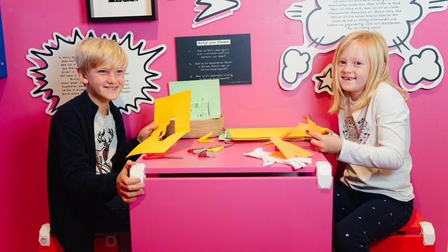 Im Bild: Ein Bub und ein Mädchen sitzen vor einer rosa Wand an einem rosa Tisch und schneiden gelbes Papier aus. 