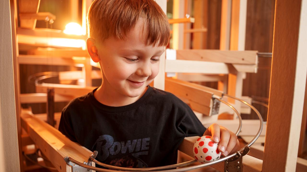 Im Bild: Ein Bub steht vor einer Kugelbahn und hält eine bunte Kugel darauf fest. 