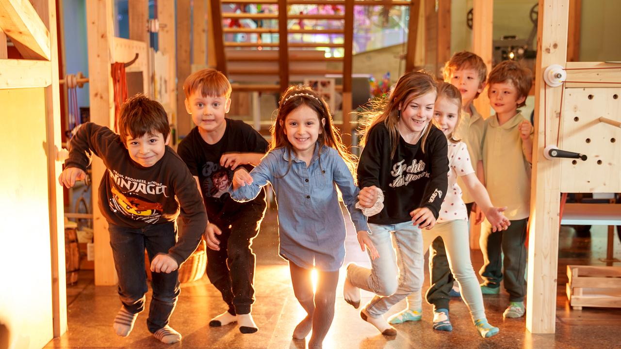 Im Bild: 5 gut gelaunte Kinder laufen durch das Kindermuseum, zwei stehen im Hintergrund. 
