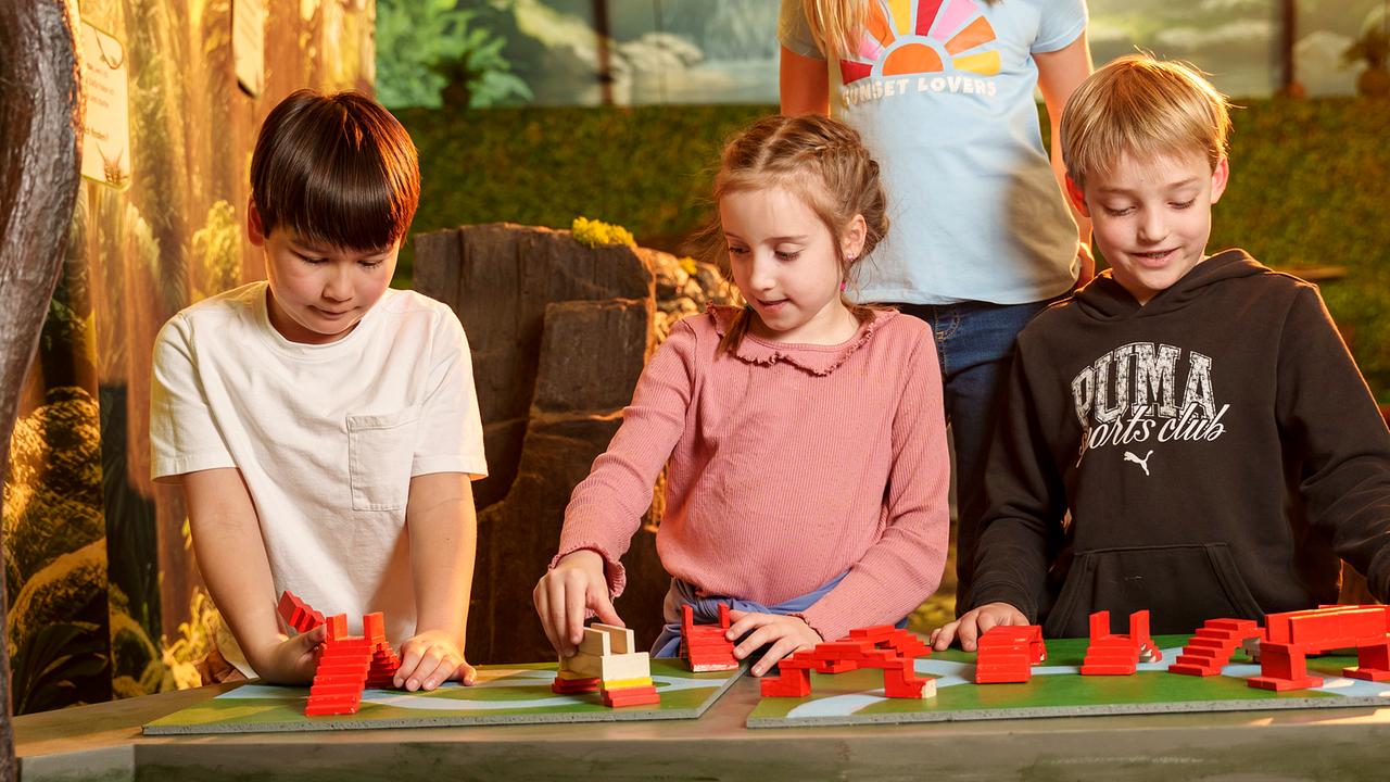 Im Bild: Zwei Buben und ein Mädchen stehen bei einem Tisch und experimentieren mit Holzformen. 