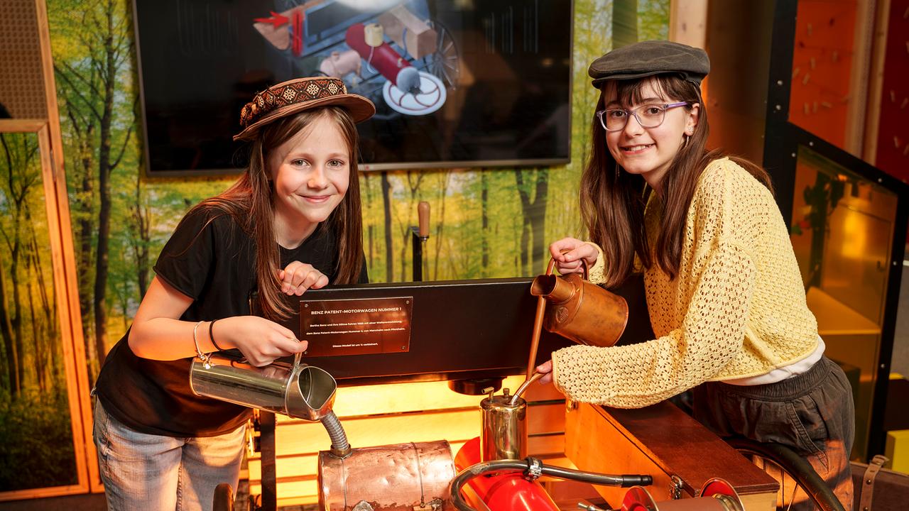 Im Bild: Zwei Mädchen stehen im Museum und gießen eine Flissigkeit in eine Maschine.