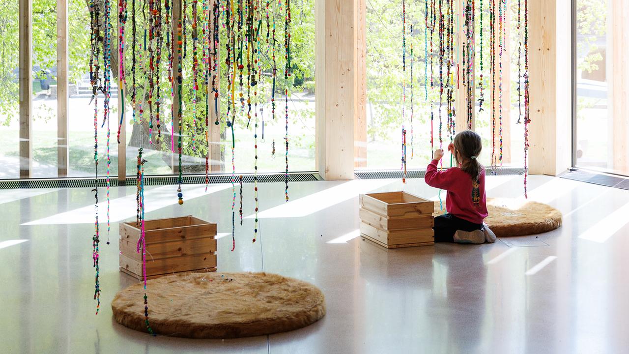 © Max Kropitz Ein Mädchen sitzt am Boden und spielt sich mit den bunten Perlenketten, die von der Decke herunter hängen.
