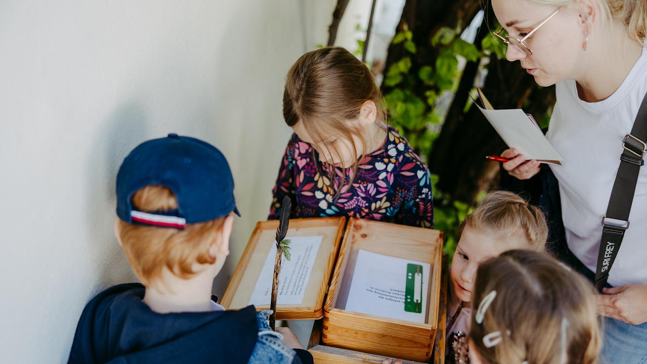 Vier Kinder und eine Frau stehen vor einer kleinen Truhe und lesen die Texte, die sich darin befinden.