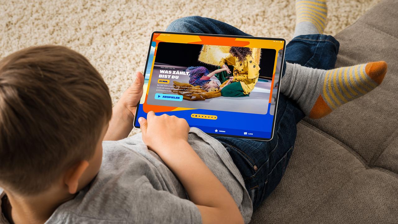 Ein Bub sitzt auf einem Sofa und schaut in ein Tablet. Auf dem Bildschirm ist die Startseite von kids.ORF.at zu sehen.