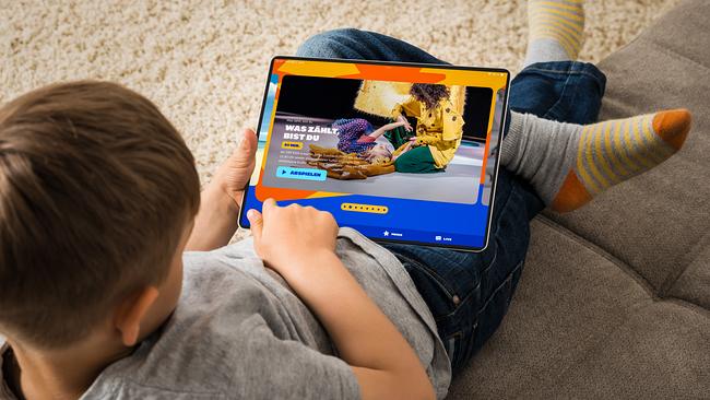 © Adobe Stock Ein Bub sitzt auf einem Sofa und schaut in ein Tablet. Auf dem Bildschirm ist die Startseite von kids.ORF.at zu sehen.