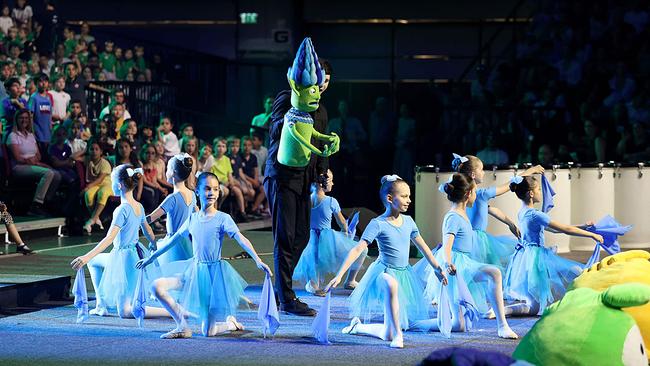 Kinder aus einer Ballettgruppe führen einen Tanz auf. Sie sind weiß gekleidet und knien am Boden, in der Mitte steht eine Grüne Figur.