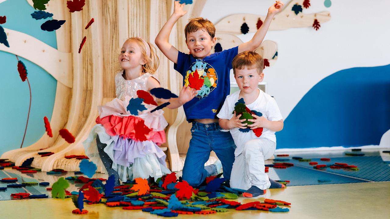 Im Bild: Drei Kinder kniehen neben einem Holbaum und spielen mit Blättern aus Filz. 