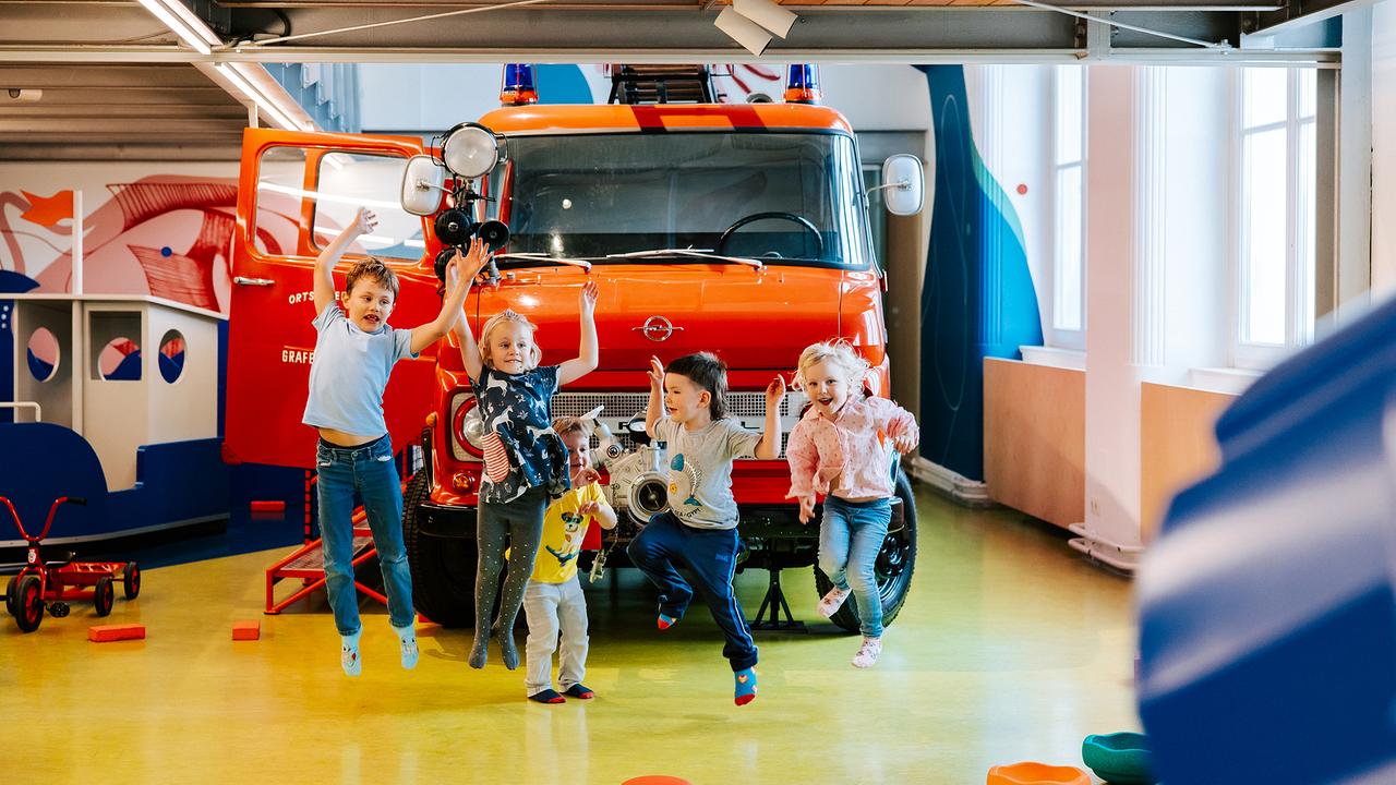 Im Bild: Fünf Kinder befinden sich im Museum und hüpfen vor einem alten Feuerwehrauto auf und ab.