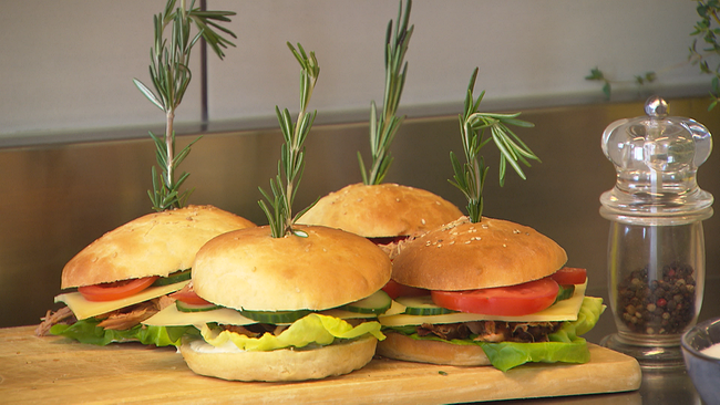 Vier fertig angerichtete Burger mit jeweils einem Rosmarienzweig dekoriert. Daneben steht eine Pfeffermühle aus Glas