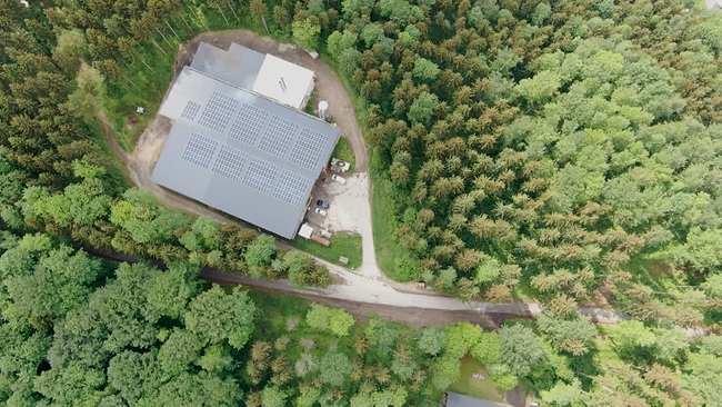 Luftbild einer Hackschnitzelanlage mit Photovoltaikanlage auf dem Dach, umgeben von Wald
