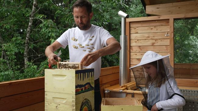 Zwei Personen an einem Bienenkorb, der Mann zeiht einen Rahmen heraus und zeigt sie einem Kind, das einen Bienenhut trägt
