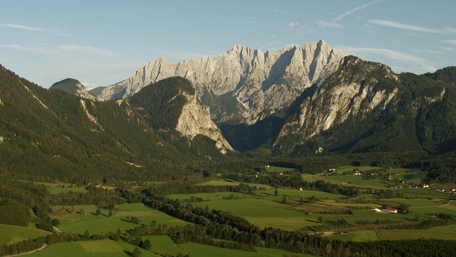 Felsiges Gebirge im Hintergrund. Davor ein Panorama aus bewaldeten Hügeln und vereinzelt gelegenen Bauernhöfen in der Landschaft