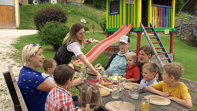 © ORF NÖ Gruppe von Erwachsenen und Kindern sitzt auf einem Tisch im Freien und bekommt gerade Essen serviert