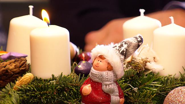© ORF NÖ Eine brennende Kerze auf einem Adventkranz, davor die Figur eines Wichtels, der die Kerze ansieht