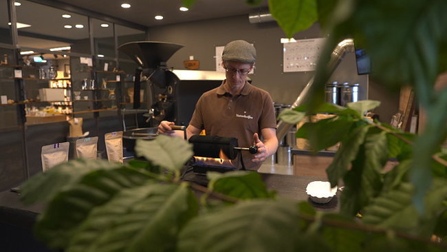 Eine Person röstet Kaffee über Feuer, im Bildvordergrund sind grüne Blätter