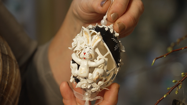 eine Person hält ein kunstvoll geschnitztes Osterei mit einem Hasenornament in die Kamera