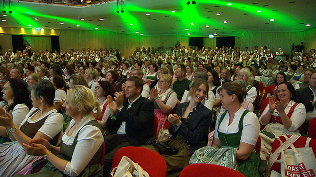 Festsaal in grünem Licht, Besucherinnen und Besucher, überwiegend in Tracht gekleidet, applaudieren