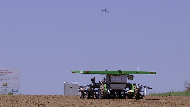 Feldroboter fährt auf einem Acker, Drohne fliegt über ihm