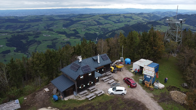 Ein Berg aus der Adlerperspektive. Im Vordergrund ist eine Schutzhütte zu sehen und Baumaterialien. Auf dem Dach der Hütte sind Photovoltaikpanele angebracht