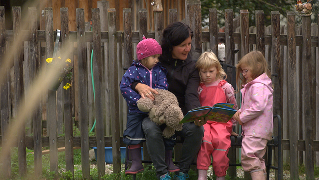 Kindergartenbetreuerin mit 3 Kleinkindern, sitzt vor Holzzaun und liest Buch vor