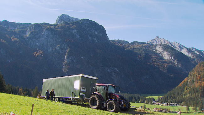 Traktor mit einem langen grünen Anhänger, der vor der Kulisse von Bergen steht. Davor zwei Menschen, die auf der Wiese am Anhänger entlang gehen