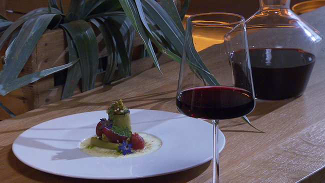 Lauchgericht auf einem weißen Teller. Daneben steht ein Glas Rotwein und eine Karaffe gefüllt ebenfalls mit Rotwein auf einem Holztisch. Dahinter als Dekoration, frische Lauchblätter