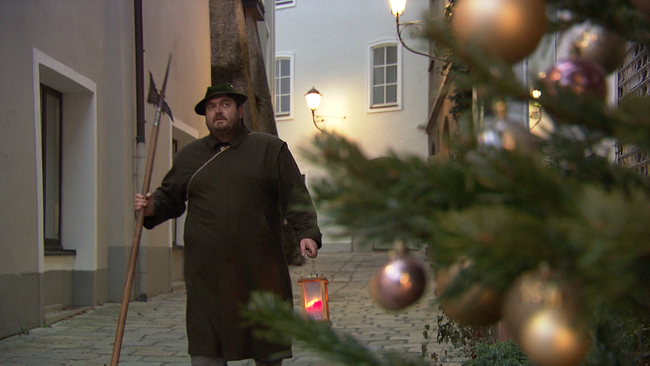 Eine Person geht mit einem Stock an dessen Ende eine Metallspitze steckt an einem geschmückten Baum in einer engen Gasse vorbei. In der andern Hand hält er eine Laterne mit einer brennenden Kerze